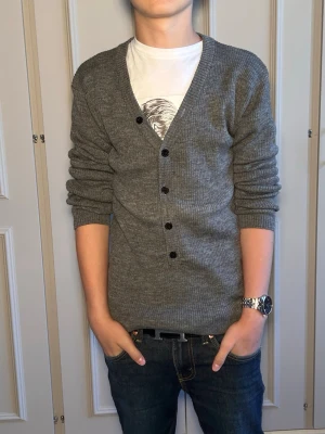 Grå ribbstickad kofta med knappar - Snygg grå ribbstickad kofta med svarta knappar och v-ringning. Långärmad modell som passar perfekt över en t-shirt. Enkel och stilren design som funkar till det mesta.