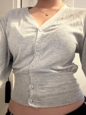 Ljusgrå ribbad kofta med knappar - Snygg ljusgrå kofta med långa ärmar och vita knappar hela vägen framtill. Modellen är figurnära med ribbad midja och v-ringad hals. Perfekt att styla med jeans eller kjol för en chill look.