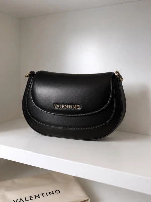 Svart handväska från Valentino - Snygg svart handväska från Valentino i slätt konstläder med guldfärgade metalldetaljer och logotyp framtill. Väskan har rundad form och stilren design, perfekt för att lyfta din outfit.