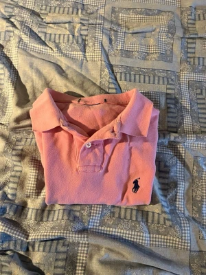 Rosa pikétröja från Ralph Lauren - Klassisk rosa pikétröja från Ralph Lauren med den ikoniska broderade loggan på bröstet. Tröjan har krage, knappar framtill och korta ärmar. Perfekt för en avslappnad men ändå stilren look. Finns ett litet litet hål nedanför loggan, men lätt att laga och inget man tänker på. Säljer då den är lite kort på mig, är 179cm. Hör av er vid frågor