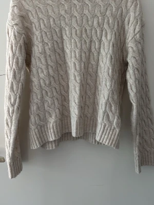 Beige kabelstickad tröja - Säljer en beige kabelstickad tröja från HM med rund hals och långa ärmar. Tröjan har ett klassiskt flätmönster och ribbade muddar vid ärmslut och nederkant. Använd fåtal gånger och är nästan i helt nytt skick. 