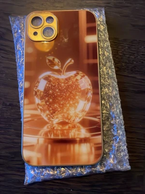 Guldigt mobilskal med äpple-motiv - Snyggt mobilskal i guld med ett glansigt 3D-äpple-motiv på baksidan. Skalet har en spegelblank finish och är gjort i hårdplast. Passar iPhone 14 Plus och har exakta utskärningar för kamera och knappar. Perfekt för dig som vill sticka ut med ett unikt och lyxigt skal.