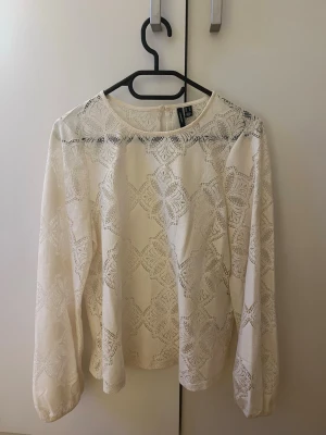 Crémefärgad blus från Vero Moda  - Snygg vit blus från Vero Moda i storlek XS. Blusen är tillverkad i ett genombrutet spetsmaterial med vackert mönster och har rund halsringning samt långa ärmar. Perfekt för dig som gillar en romantisk och stilren look.