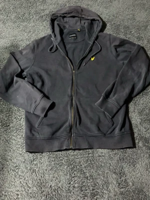Svart hoodie från Lyle & Scott - Svart hoodie med dragkedja från Lyle & Scott. Klassisk design med huva och snörning, samt gul broderad logga på bröstet. Mjuk bomullskänsla och ribbade muddar vid ärmslut och nederkant. Perfekt för en avslappnad streetstyle.