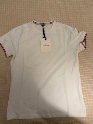 Vit Moncler t-shirt med logga - Vit t-shirt från Moncler med klassisk rund hals och korta ärmar. T-shirten har Moncler-logga på ärmen och detaljer i rött och blått vid halsringning och ärmslut. Tillverkad i mjuk bomull för en skön känsla.   Oanvänd i storlek M, liten blå fläck under armen, går säkert få bort ej testat, super fin pasform slim och skön kvalite