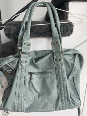 Ljusblå handväska med flätade detaljer - Snygg ljusblå handväska i mjukt skinnliknande material med dubbla handtag och flätade detaljer framtill. Väskan har en praktisk ytterficka med dragkedja och dekorativa metalldetaljer. Perfekt för dig som vill ha en stilren och rymlig väska.