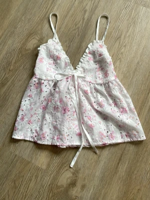 Vit blommig linnetopp med spetsdetaljer - Supersöt vit linnetopp med rosa blommigt mönster, broderade hål och smala axelband. Toppen har v-ringning, volangkant och knytband framtill. Perfekt för varma dagar och ger en romantisk vibe.