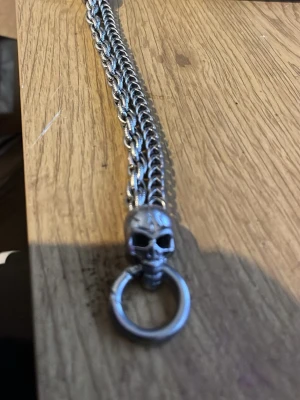 Kedja med dödskallehänge i rostfritt stål  - Cool armband i silverfärgad metall med ett detaljerat dödskallehänge som håller en ring. Kedjan har en robust och flätad design, perfekt för dig som gillar en edgy och alternativ stil.