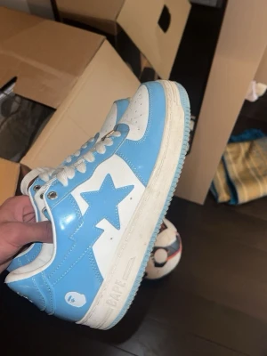 BAPE STA blåvita sneakers - Säljer ett par BAPE STA sneakers i blått och vitt med klassisk stjärndesign på sidan. Skorna har vita snören, perforerad tåbox och BAPE-logga på hälen. Tillverkade i glansigt syntetmaterial och har en låg silhuett som ger en clean streetwear-look.