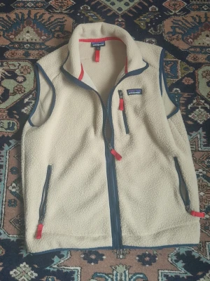 Patagonia Väst - Säljer en beige fleeceväst från Patagonia med blå dragkedjor och röda detaljer. Västen har hög krage, tre fickor med dragkedja och är tillverkad i mjukt fleece-material. Perfekt för lager-på-lager och outdoor-vibe. I mycket fint skick, helt utan defekter  