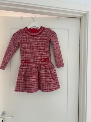 Röd och vit rutig klänning - Snygg röd och vit rutig klänning för barn med lång ärm och rund hals. Klänningen har markerad midja och utställd kjol, samt dekorativa detaljer vid midjan. Perfekt för dig som gillar en klassisk men trendig look. Storlek 122 passar en 6-7 åring.
