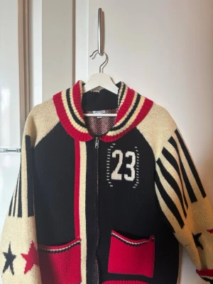 Stickad ziptröja Jaded London, svart/röd - Cool stickad ziptröja från Jaded London i svart, rött och beige med nummer 23 på bröstet, randiga ärmar och stjärndetaljer. Två röda fickor framtill och bred krage med ränder. Perfekt för dig som gillar streetstyle och unika plagg.