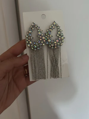 Glittriga statement örhängen med strass - Säljer ett par stora statement örhängen från H&M med droppformad design, täckta av glittrande strass i regnbågsfärger och långa silverfärgade kedjor som hänger ner. Perfekta för att sticka ut och ge extra bling till din look.