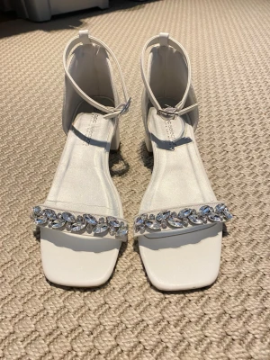 Vita sandaler med strass och blockklack - Stilrena vita sandaler från boohoo med fyrkantig tå, bred rem framtill dekorerad med gnistrande strass och justerbart ankelband. Blockklack ger stabilitet och en modern look. Perfekta för dig som vill ha något extra till outfiten.