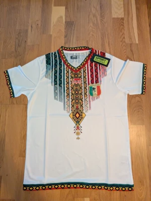 Mali fotbollströja -  Helt ny Vit fotbollströja med korta ärmar och färgglada, etno-inspirerade mönster i rött, grönt, gult och svart längs halsringning, ärmslut och nederkant. Tryckt emblem på bröstet och V-ringad krage. Perfekt för dig som gillar unika plagg med mycket färg.