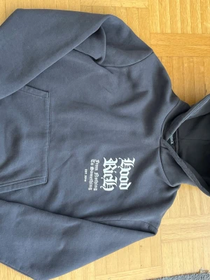 Mörkgrå Hoodrich hoodie med tryck s/m - Snygg mörkgrå hoodie från Hoodrich med vit text på bröstet och stort grafiskt tryck på ryggen. Klassisk känguruficka framtill och huva. Perfekt för dig som gillar streetwear och vill sticka ut med coola detaljer.