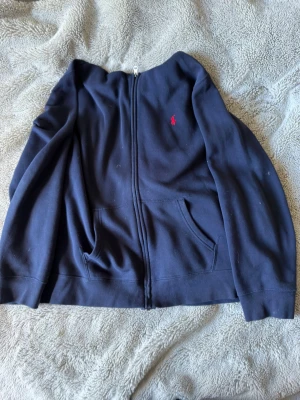 Mörkblå hoodie från Ralph Lauren - Mörkblå hoodie med dragkedja från Ralph Lauren. Klassisk design med huva, två fickor framtill och röd broderad logga på bröstet. Tillverkad i mjukt bomullsmaterial, perfekt för en avslappnad och stilren look.
