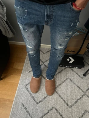 Blå slitna slim jeans - Säljer ett par blå slim jeans med slitningar och hål på framsidan. Jeansen har klassisk femficksdesign och smal passform hela vägen ner. Perfekta för en avslappnad och trendig streetstil.
