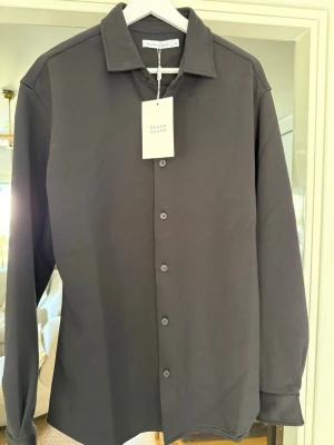 Svart skjorta från Brand Island XL - Stilren svart skjorta från Brand Island i storlek XL. Skjortan har klassisk krage, knäppning framtill och långa ärmar. Perfekt för dig som gillar en enkel och clean look. Materialet känns mjukt och bekvämt mot huden.
