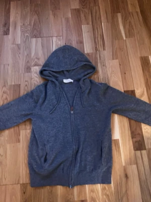 Mörkgrå zip-hoodie med huva - En enkel och stilren mörkgrå hoodie med dragkedja framtill och två fickor. Hoodien har en klassisk huva med dragsko och är tillverkad i ett mjukt, stickat material. Perfekt för en avslappnad look. Tröjan har två små hål men de syns knappt när du har på dig den
