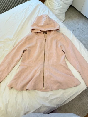 Ljusrosa hoodie med dragkedja - Mysig ljusrosa hoodie med dragkedja framtill och huva. Modellen har en lätt utsvängd passform nedtill och långa ärmar. Materialet är mjukt och ser ut att vara bomullsblandning. 