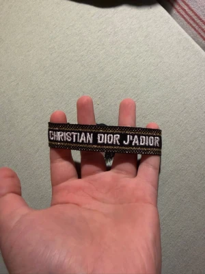  svart armband - Snyggt svart tygarmband ' broderad i vitt. Armbandet har guldfärgade detaljer och justerbar knytning med fransar. Perfekt accessoar för att lyfta din outfit.