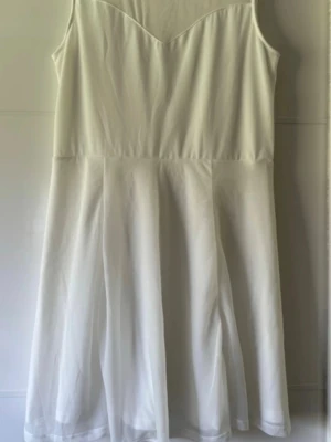 Vit ärmlös kortklänning med mesh - Snygg vit kortklänning med ärmlös design och meshdetalj upptill. Klänningen har en lätt utställd kjol och rund halsringning med transparent mesh över bröstet. Perfekt för sommarens alla tillfällen. Passar till studenten.