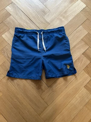 U.S Polo Assn - Blå shorts från U.S Polo Assn med vit snörning i midjan och gul broderad logga på benet. Klassisk modell med elastisk midja och bakficka. Perfekta för sommaren och chill dagar.