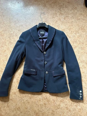Mörkblå kavaj från Pikeur - Snygg mörkblå kavaj från Pikeur med tre silverfärgade knappar framtill och dekorativa knappar vid ärmsluten. Kavajen har klassisk krage, två fickor med lock och är helfodrad med glansigt tyg. Perfekt för en stilren look.( jag vill igentligen absolut inte sälja den här men tyvärr är den för liten för mig så bättre att den får glädja någon annan)
