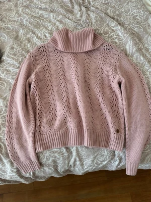 Rosa stickad polotröja med hålmönster - Mysig rosa stickad polotröja med dekorativt hålmönster och bred ribbad krage. Tröjan har långa ärmar och ribbade muddar vid ärmslut och nederkant. Perfekt för kyliga dagar och enkel att styla till jeans eller kjol.