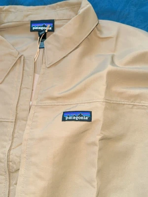 Beige vindjacka från Patagonia - Snygg beige vindjacka från Patagonia med dragkedja framtill, klassisk krage och två sidofickor. Jackan har elastiska muddar nedtill och logotyp på bröstet. Perfekt för blåsiga dagar och enkel att matcha med olika outfits.