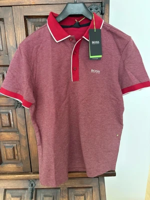 Röd pikétopp från BOSS Athleisure - Röd pikétopp från BOSS Athleisure med vit kant på kragen och knäppning framtill. Regular fit och kortärmad modell med diskret rutigt mönster och broderad logga på bröstet. Perfekt för en sportig och stilren look. Aldrig använd. 