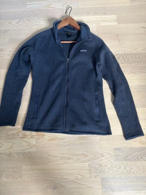 Mörkblå fleecejacka från Patagonia - Mysig mörkblå fleecejacka från Patagonia med hel dragkedja och två fickor framtill. Jackan har hög krage och broderad logga på bröstet. Perfekt för lager-på-lager och chill dagar.