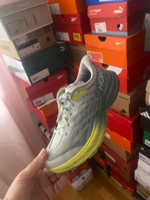 Hoka grå-gula sneakers - Snygga sneakers från Hoka i grått meshmaterial med gula detaljer och en chunky, gulgrå sula. Skorna har en sportig vibe och är perfekta för dig som gillar streetstyle och komfort. Klassisk snörning och Hoka-logga på sidan.