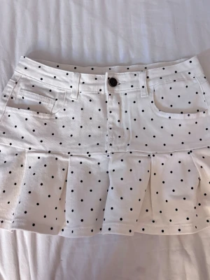 Vit prickig kortkjol med volang - Superfin vit kortkjol med svarta prickar och volang nertill. Kjolen har fickor fram, bälteshällor och knäppning med knapp och dragkedja. Perfekt för en trendig och lekfull look.