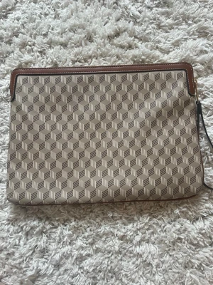 Beige datorfodral med grafiskt mönster - Dator fodral i beige och brunt med grafiskt kubmönster och bruna läderdetaljer. Väskan har en avtagbar handledsrem i brunt läder och dragkedja upptill. Perfekt för att bära det viktigaste med stil.