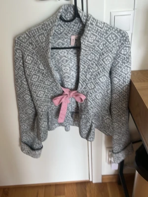 Grå mönstrad kofta med rosa rosett - Superfin grå kofta med vitt geometriskt mönster och en söt rosa rosett framtill. Koftan har lång ärm, öppen front och är stickad i mjukt material. Perfekt att slänga över en topp för en mysig look.