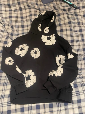 Svart hoodie med vita blommor - Svart hoodie med huva och stora vita blommönster tryckta över hela plagget. Hoodien har ribbade muddar vid ärmslut och nederkant. Perfekt för dig som gillar streetwear och vill sticka ut med en cool design.
