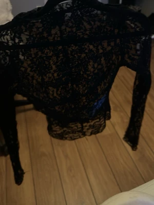 Svart spetsblus med lång ärm - Säljer en svart, transparent spetsblus med lång ärm och hög hals. Blusen har ett detaljerat blommönster i spets och är figurnära i modellen. Perfekt för dig som vill ha en edgy och trendig look.