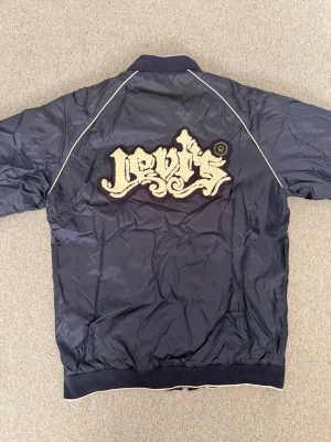 Svart bomberjacka från Levi's - Snygg svart bomberjacka från Levi's med vit piping längs axlarna och stor broderad logga i vitt på ryggen. Hör av dig vid funderingar och behov av fler bilder!🤗