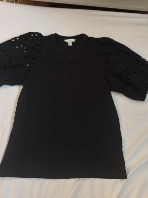 Svart topp med puffärmar från H&M!! - Svart topp från H&M med snygga puffärmar i broderad bomull och hålmönster. Klassisk rund halsringning och rak passform. Perfekt för dig som vill ha en enkel men ändå trendig look.