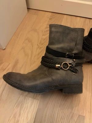 gråa biker ankle boots - ankle boots med låg klack. tunt innerfoder i fuskpäls. snygg dekoration - rem med spänne, svarta flätor och metall ringar. zipper på insidan, märke marco tozzi och skorna är gjort av syntetläder av hög kvalitet. använt en del men inga defekter som märkts av. 