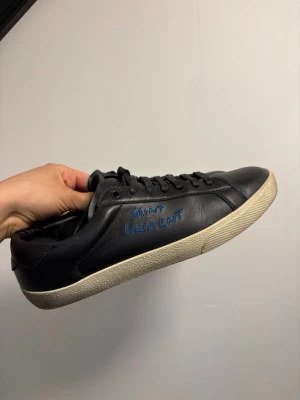Saint Laurent court sneakers svarta  - Svarta sneakers från Saint Laurent i mjukt läder med blå broderad logga på sidan. Klassisk låg modell med svarta skosnören och vit sula. Perfekta för dig som gillar stilrena och exklusiva sneakers med en touch av lyx.