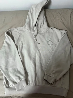 Beige hoodie med smiley-tryck - Snygg beige hoodie från Just Be Kind med huva och dragsko. På bröstet finns ett litet tryck med text och på ryggen ett stort svart smiley-tryck som blinkar. Mjuk bomullskänsla och ribbade muddar vid ärmar och midja.