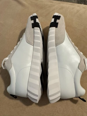 Vita och beige sneakers från Hogan - Snygga sneakers från Hogan i vitt läder med beige mockadetaljer och en chunky, randig sula. Klassisk låg modell med svart H-logga på tån. Perfekta för dig som vill ha en clean och modern look.