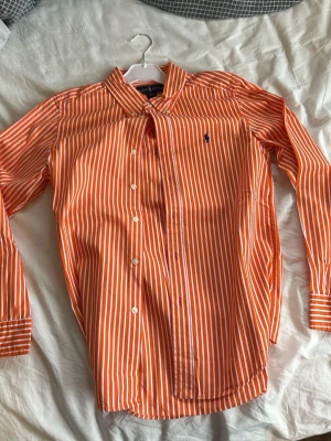 Orange- och vitrandig skjorta Ralph Lauren - Snygg orange skjorta med vita ränder från Ralph Lauren. Klassisk krage, knäppning framtill och långärmad modell. Ikonisk logga broderad på bröstet. Skjortan är i bomull och har en fräsch, ungdomlig stil.