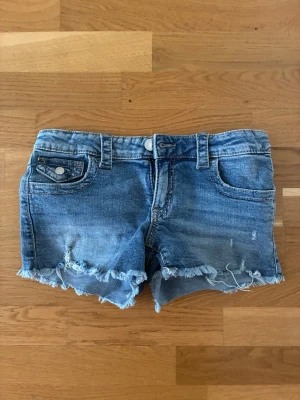 Blå jeansshorts med fransig kant - Säljer ett par blå jeansshorts från Gina Young storlek 158  med fransig rå kant och klassisk femficksdesign. Shortsen har bälteshällor, knappgylf och slitna detaljer för en avslappnad look. Perfekta för varma dagar och enkel att matcha med olika toppar.