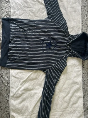 Randig hoodie från Converse - Mörkblå hoodie från Converse med vita vertikala ränder och tryckt logga på bröstet. Klassisk huva med dragsko och känguruficka framtill. Perfekt för en avslappnad streetstil.