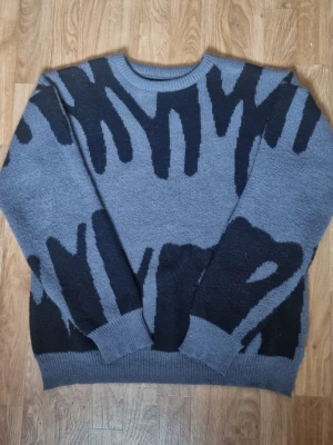 Grå stickad tröja med svart mönster - Cool grå stickad tröja med abstrakt svart mönster över hela plagget. Tröjan har rund halsringning, långa ärmar och ribbade muddar vid ärmslut och nederkant. Perfekt för dig som gillar unika stickade plagg med streetkänsla.