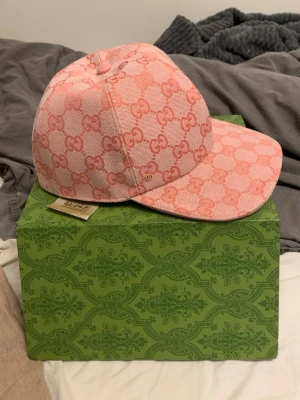 Rosa keps från Gucci med GG-mönster - Säljer en snygg rosa keps från Gucci med klassiskt GG-monogram i ljusrosa och mörkare rosa nyanser. Kepsen har böjd skärm och är tillverkad i ett vävt tyg med exklusiv känsla. Perfekt för dig som vill sticka ut med en lyxig accessoar.